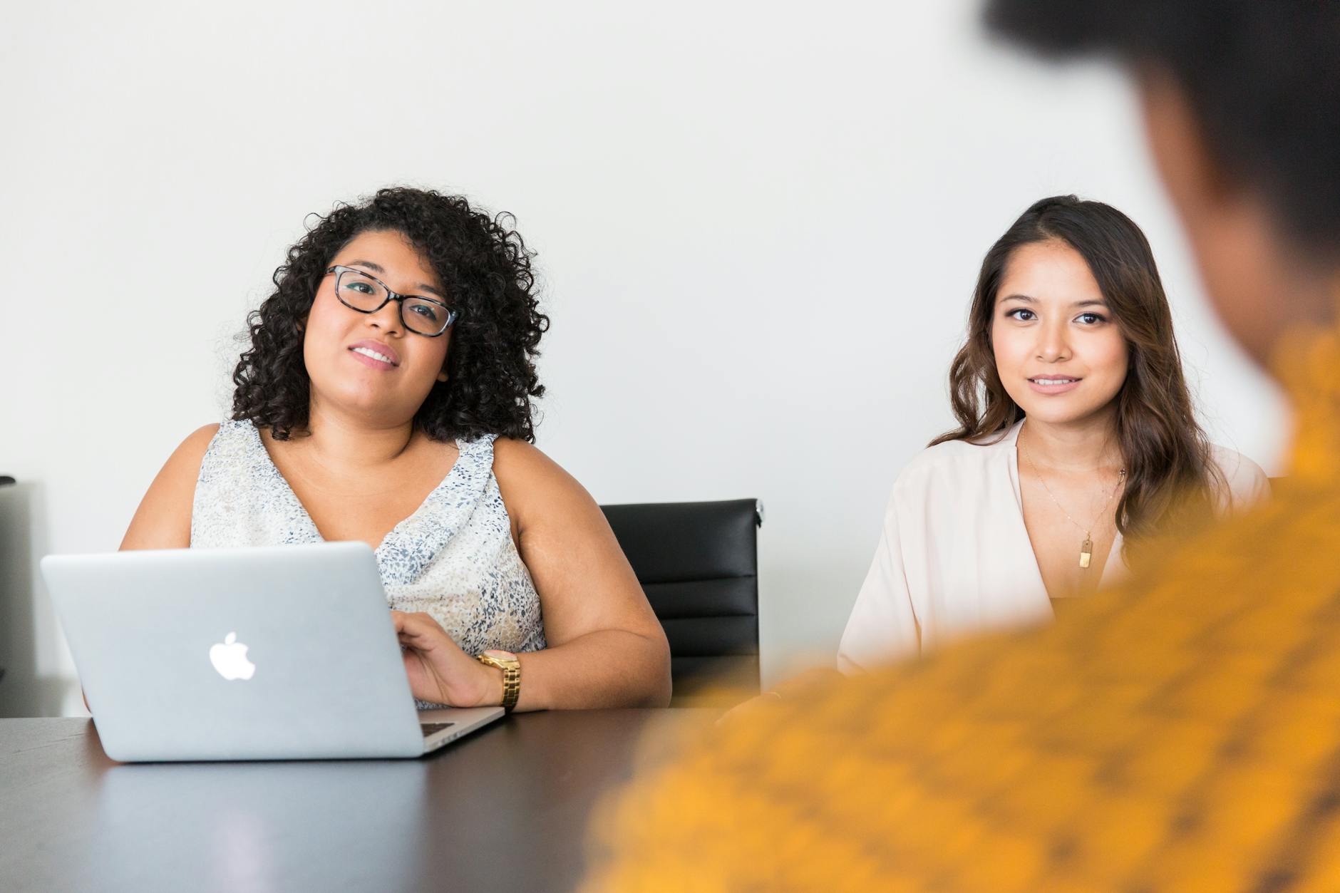 Two women sitting at a table, one with a laptop, speaking with someone in the foreground. 