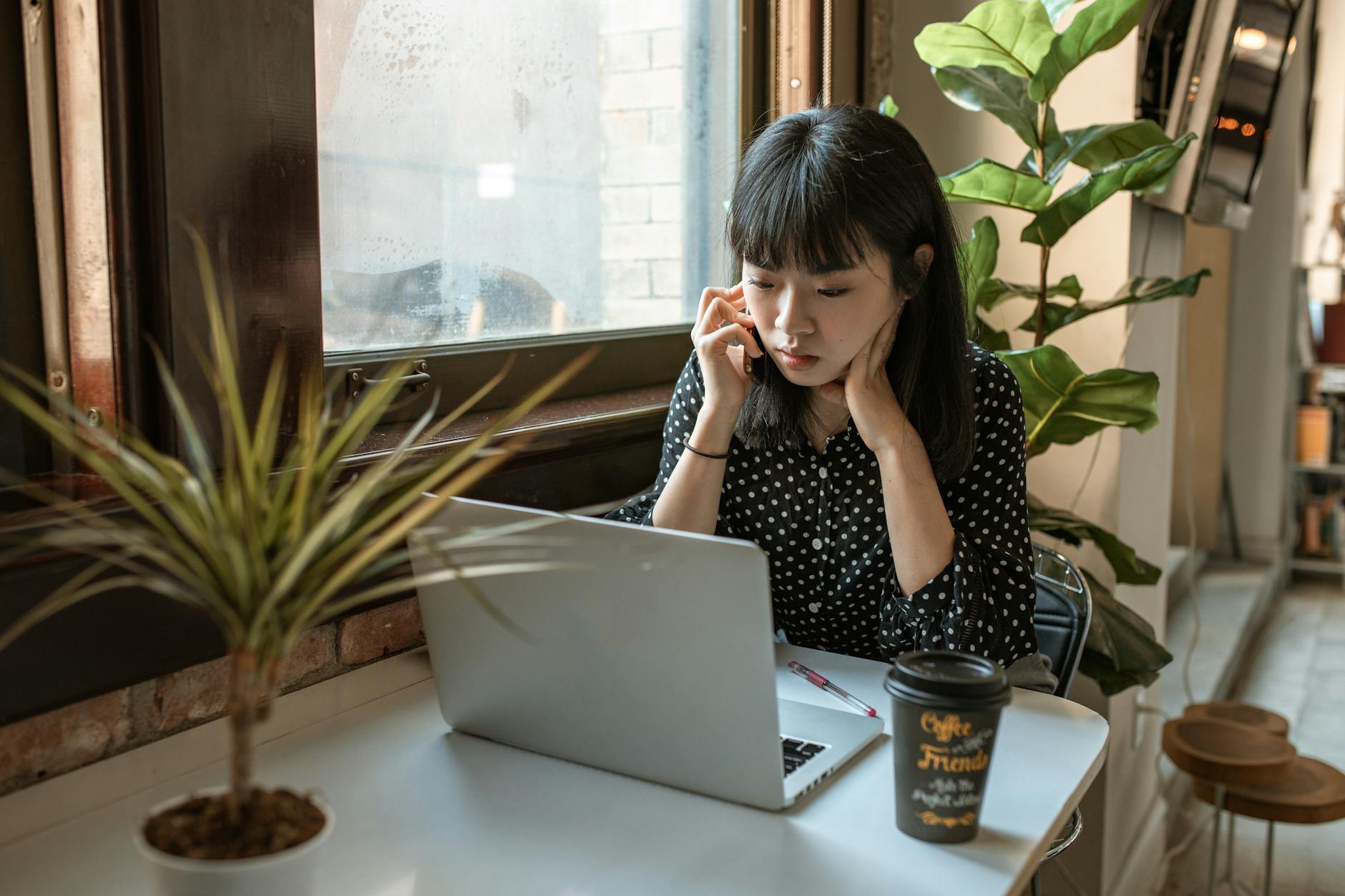 Young woman sitting at a table with a plant and a cup of coffee. Woman is on cellphone looking at laptop.