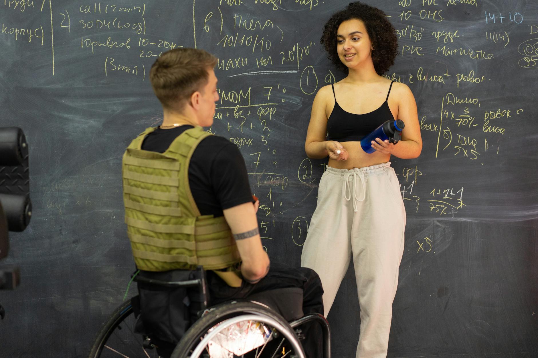 Two people in front of a chalkboard with writing on it. One is a woman in gym clothes, speaking with a man in a wheelchair wearing a military vest.